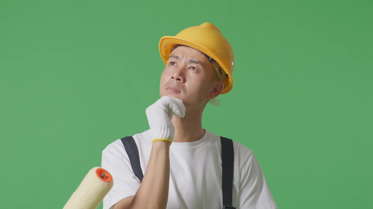 Close Up Of Asian Man Painter Wearing Safety Helmet Thinking About Something And Looking Around While Standing In The Green Screen Background Studio