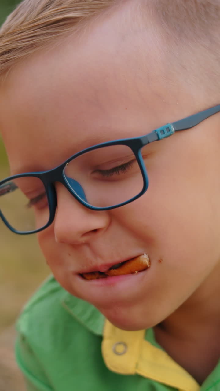 niño de camisa verde se mete todo su bocadillo en la boca mientras su hermana de camisa rosa disfruta de la suya más suavemente al aire libre en el campo abierto, ambos sentados con una canasta de picnic a su lado