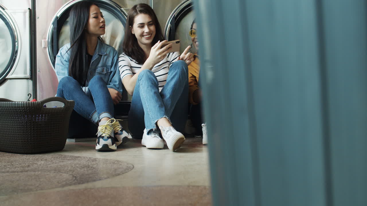 Mixed Races Girls Friends Sitting On Floor With Basket Of Dirty Clothes And Watching Video On Smartphone While Washing Machines Working