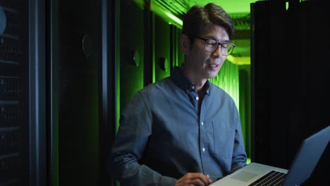 smiling asian male it technician using laptop checking computer server