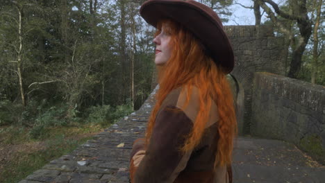 hermosa mujer pelirroja en un puente de piedra en el bosque inglés mirando a la distancia