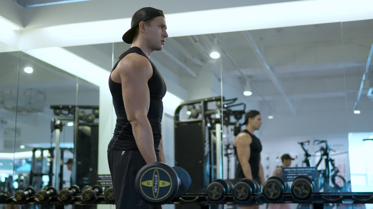 체육관 에서 비 ⁇ 스 곱슬 운동 을 하는 몸매 좋은 사람 은 거울  ⁇  에 서서 무게 를 들고 곱슬 바 를 들어 올린다