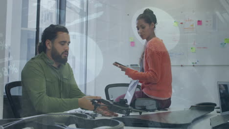 Animation-of-infographic-interface-over-diverse-man-showing-drone-model-to-coworker-in-office