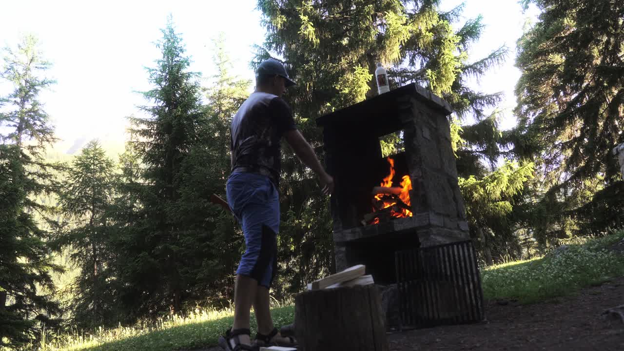 Man tending fire at an outdoor stone grill in slow motion, surrounded by nature
