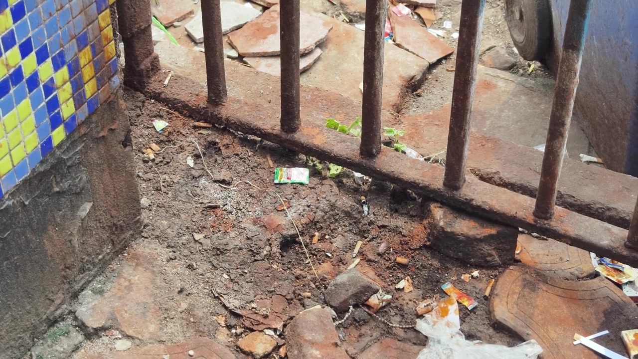 Un primo piano di rifiuti di plastica, sputo di tabacco e mozziconi di sigaretta sparsi su terreno sterrato vicino a una recinzione metallica arrugginita a Calcutta, evidenziando problemi ambientali urbani e inquinamento