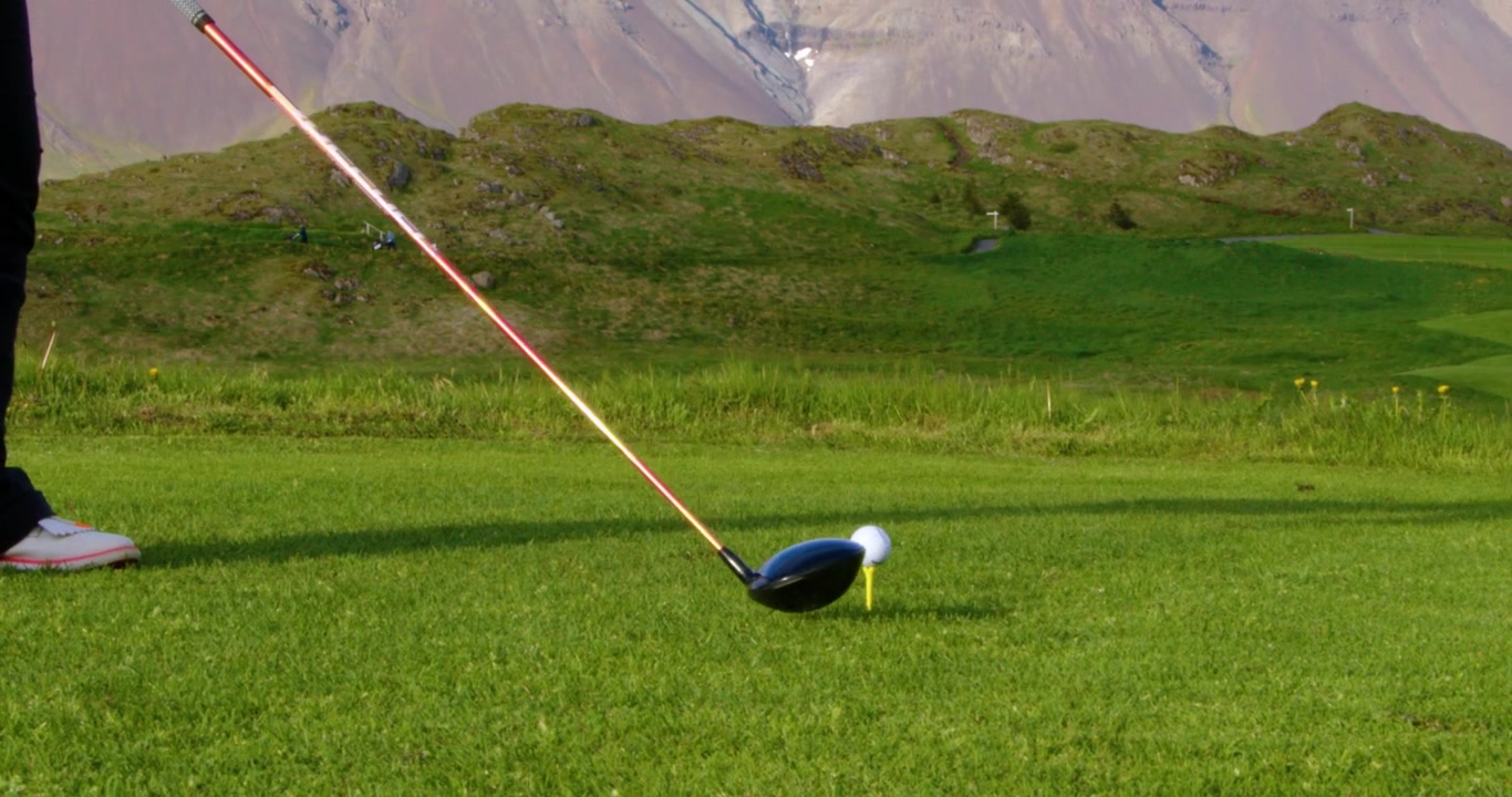 Güzel, yeşil bir sahada kadın bir golfçü driver sopasıyla başlangıç vuruşu yapmaya hazırlanıyor. Arka planda açık bir gökyüzünün altında etkileyici bir dağ silsilesi yükseliyor.
