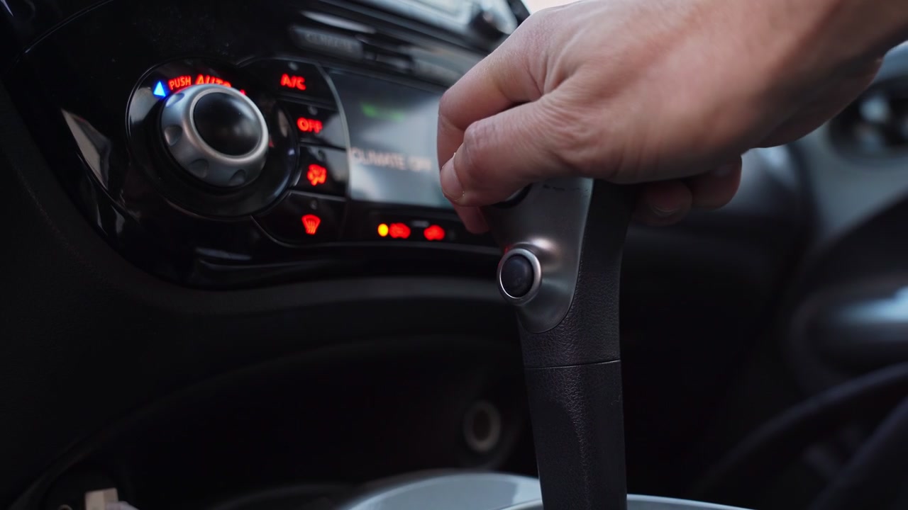 モダンな車内で、ドライバーがオートマチックギアセレクターを操作する手のクローズアップショット。ダッシュボードのエアコン操作部が、浅い被写界深度でぼやけた背景を照らしている。