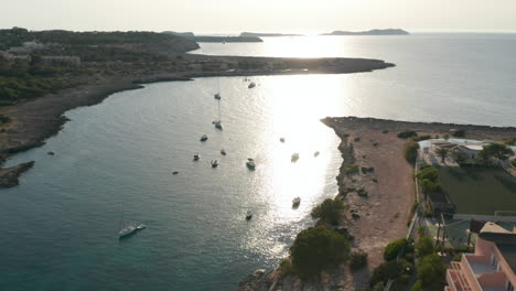 Aerial-view-of-boats-parked-in-a-silent-and-calm-sea-with-empty-resort-at-shore-with-a-football-ground-and-greenery-in-Ibiza-in-Spain