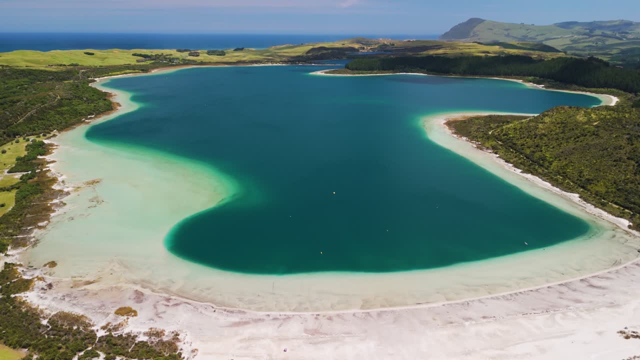 Ascending aerial pullback revealing pristine Kai Iwi Lakes with turquoise water, white sand beaches, and rolling hills under sunny New Zealand sky