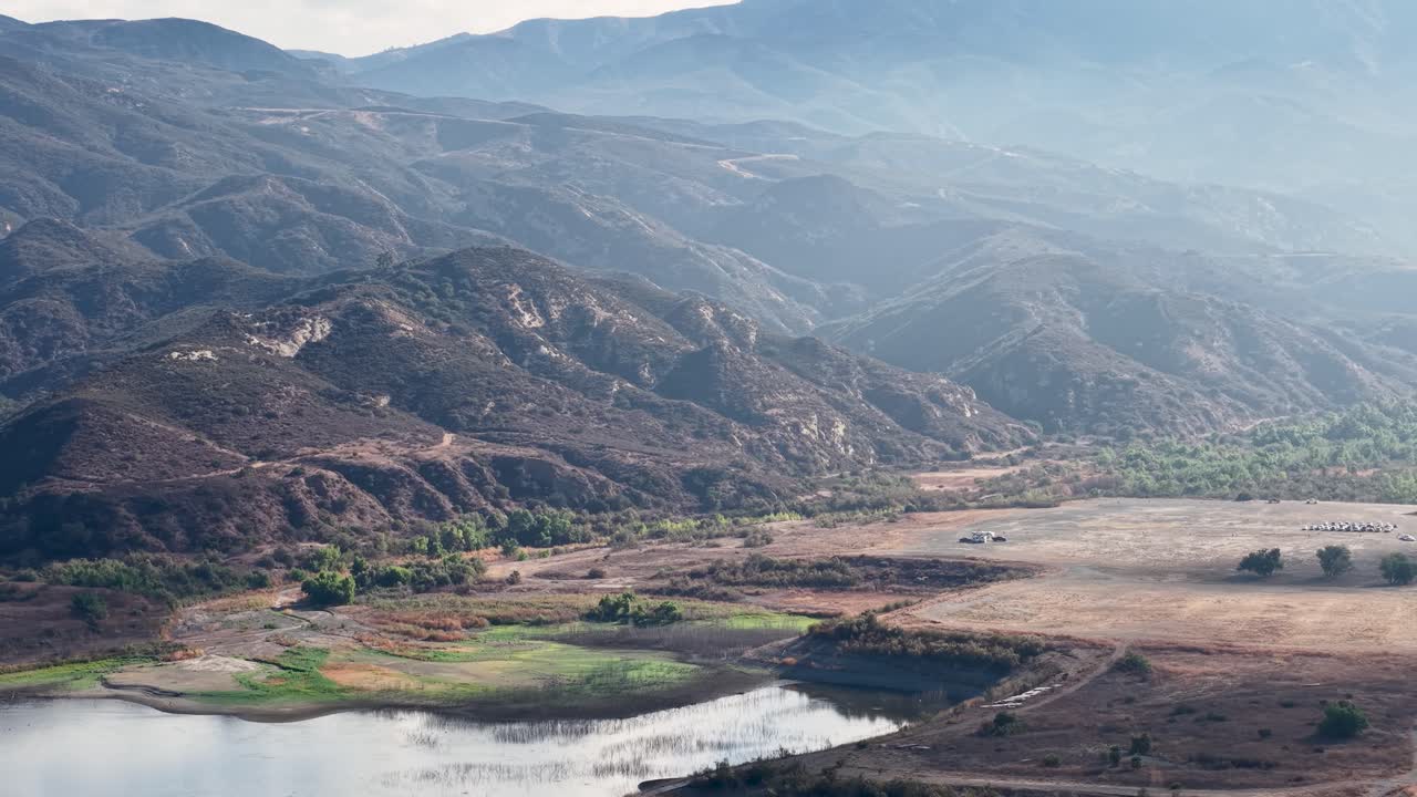 Vista aerea cinematografica sopra il Black Star Canyon in California, che mette in risalto il letto asciutto e tortuoso di un fiume, una zona di campi verdi e uno sfondo montuoso avvolto in foschia e luce solare stratificate