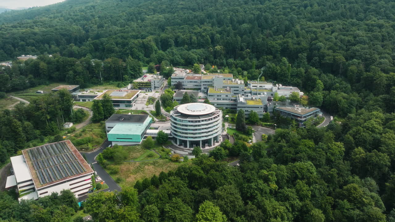 A drone performs an orbiting movement while rising and retreating above the DKFZ research campus, revealing the round building, green roofs and surrounding forest