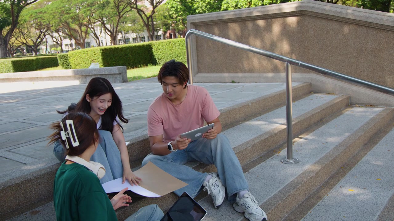 Three students sit on outdoor concrete stairs discussing project work using tablet and paper documents.