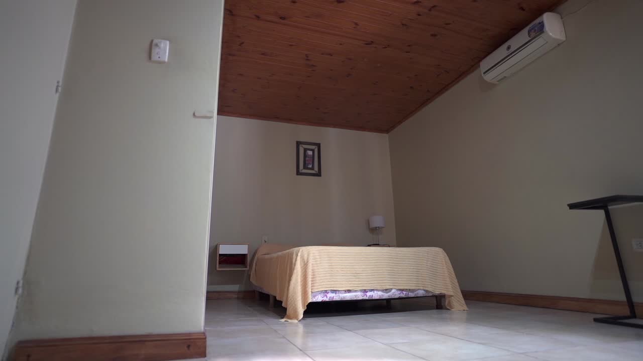 Wide angle clip shows Airbnb bedroom’s wooden ceiling, two person bed, and minimalist wall painting, with air conditioner unit for comfort. Ideal for rental listings.