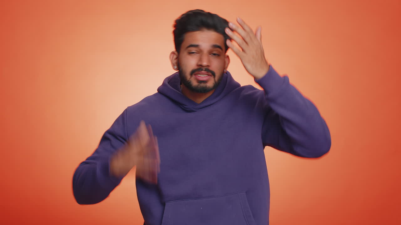 un joven parece frustrado con una expresión triste y decepcionado. está vestido con una sudadera púrpura y está de pie frente a un fondo naranja.