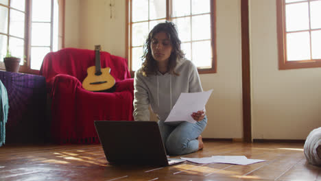 Mujer-De-Raza-Mixta-Trabajando-En-Casa,-Arrodillada-En-El-Suelo-Usando-Una-Computadora-Portátil-Y-Sosteniendo-Papeleo-En-La-Cabaña