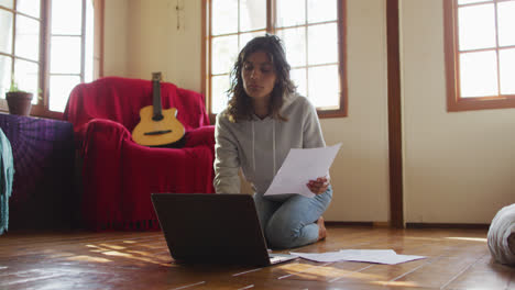 mujer de raza mixta trabajando en casa, arrodillada en el suelo usando una computadora portátil y sosteniendo papeleo en la cabaña
