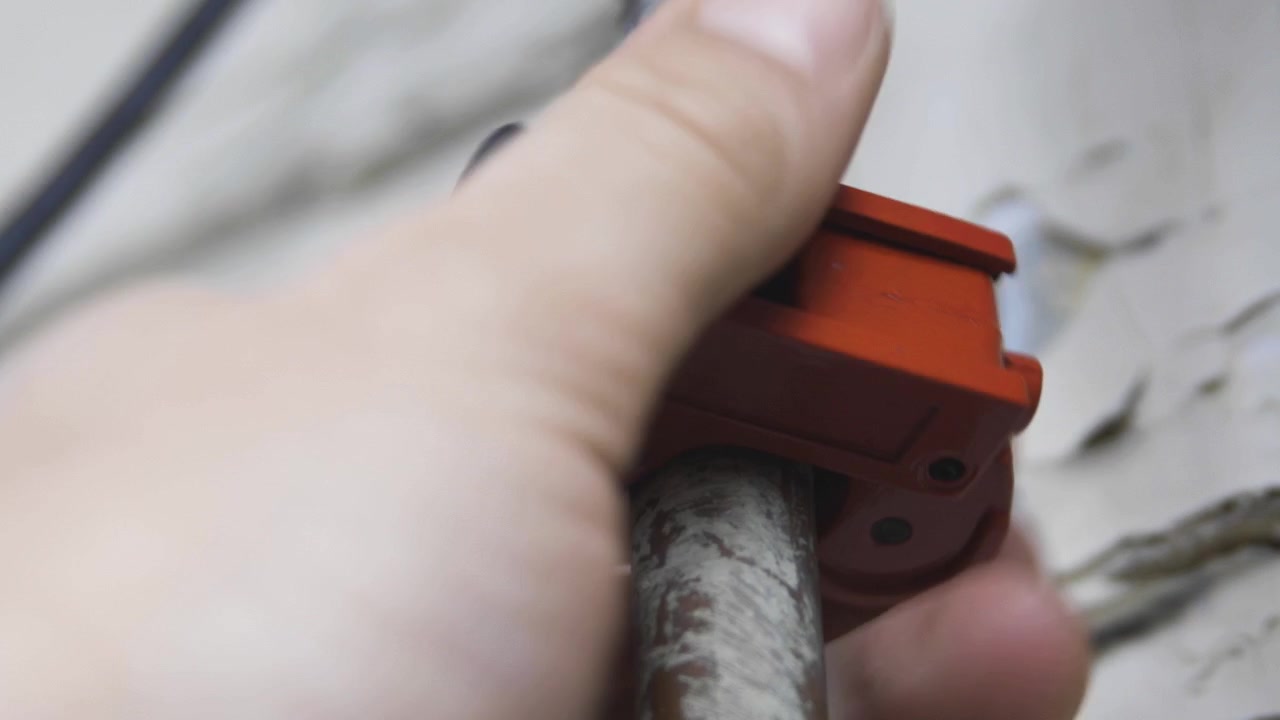 Male hand turns an orange pipecutter around an old copper pipe during repair. Low angle, closeup.