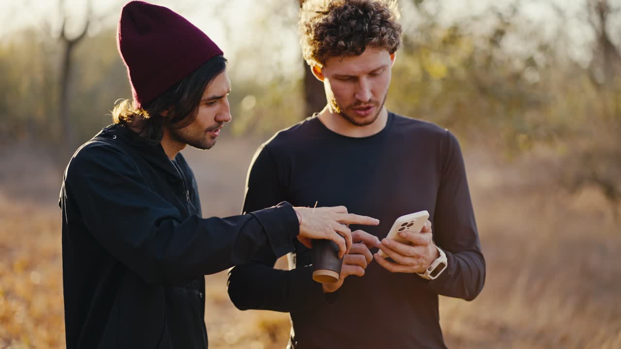 Close-up shot of two brunette athlete friends in a black sports uniform walking and talking while looking at something on the smartphone screen during their walk along an earthly path in a sunny autumn forest in the morning