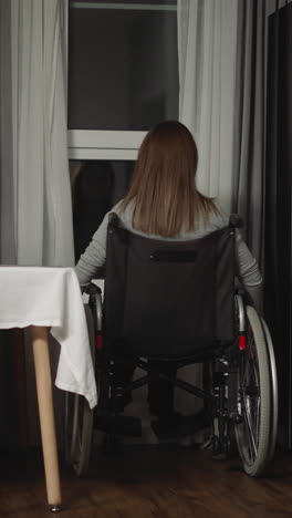 housewife moves wheelchair closer to look out small window in kitchen. woman with muscular dystrophy sits at home in evening feeling lonely backside view