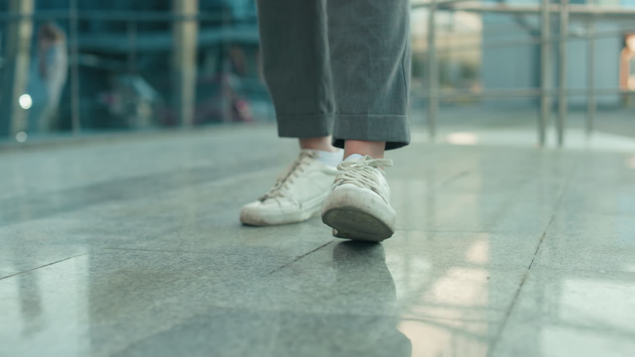 Lower angle view of individual wearing white sneakers and gray trousers dancing on sleek polished tile surface with modern glass building and metal railing in background