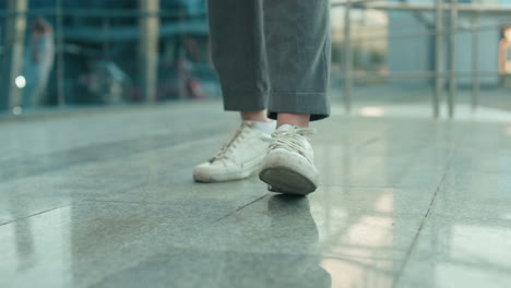 lower angle view of individual wearing white sneakers and gray trousers dancing on sleek polished tile surface with modern glass building and metal railing in background