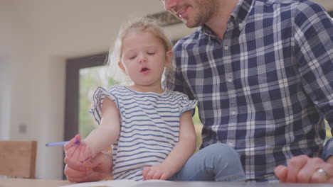 father at home at kitchen counter helping young daughter to draw picture in book - shot in slow motion