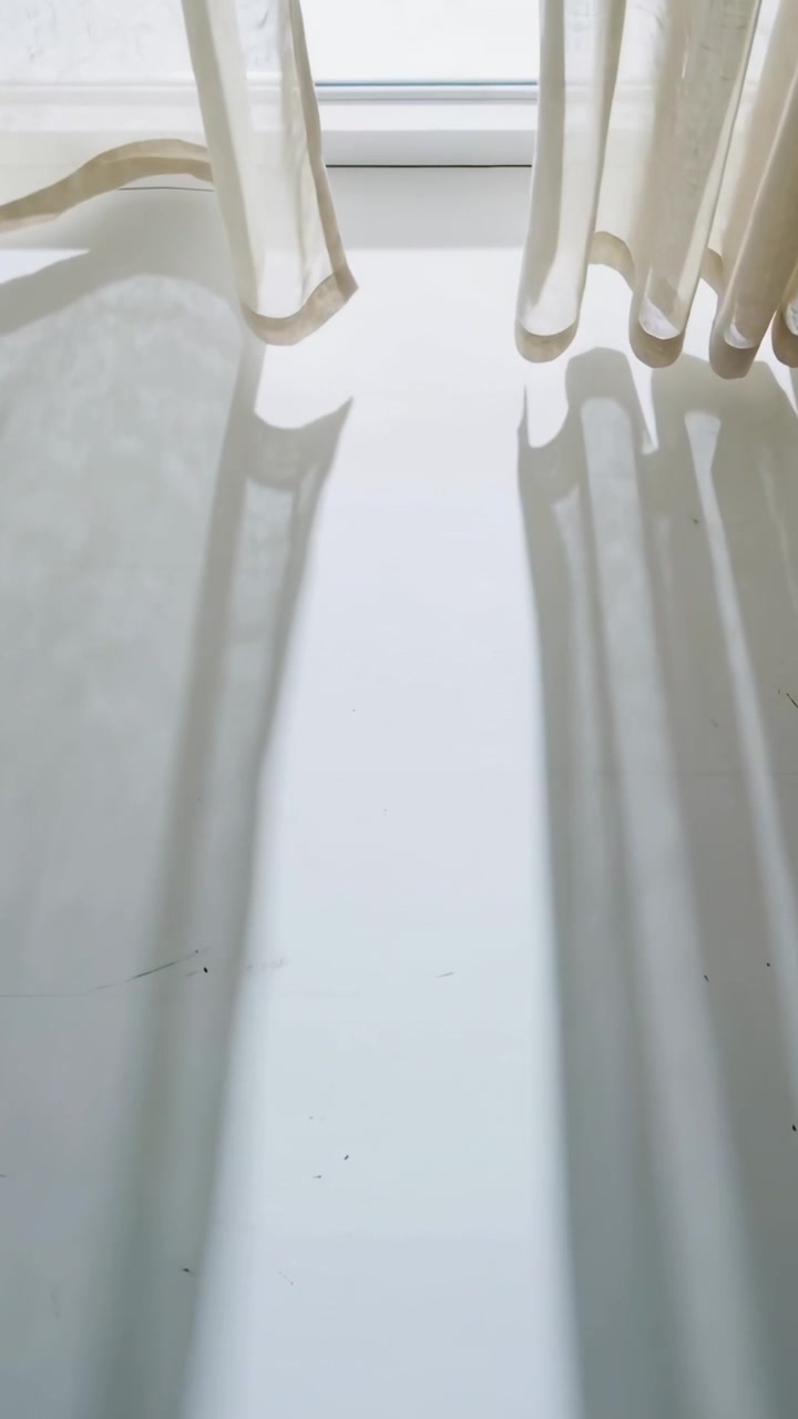 Video verticale: Raggi di sole che filtrano attraverso tende velate che si muovono alla finestra, proiettando ombre sul pavimento.