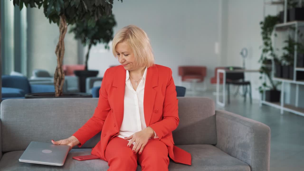 빨간색 비즈니스 슈트를 입은 노인 여성의 초상화, 비즈니스 센터의 소파에 앉아, 노트북을 닫고, 일을 끝내고, 깊은 호흡으로, 그녀는 머리에 손을  ⁇ 습니다.