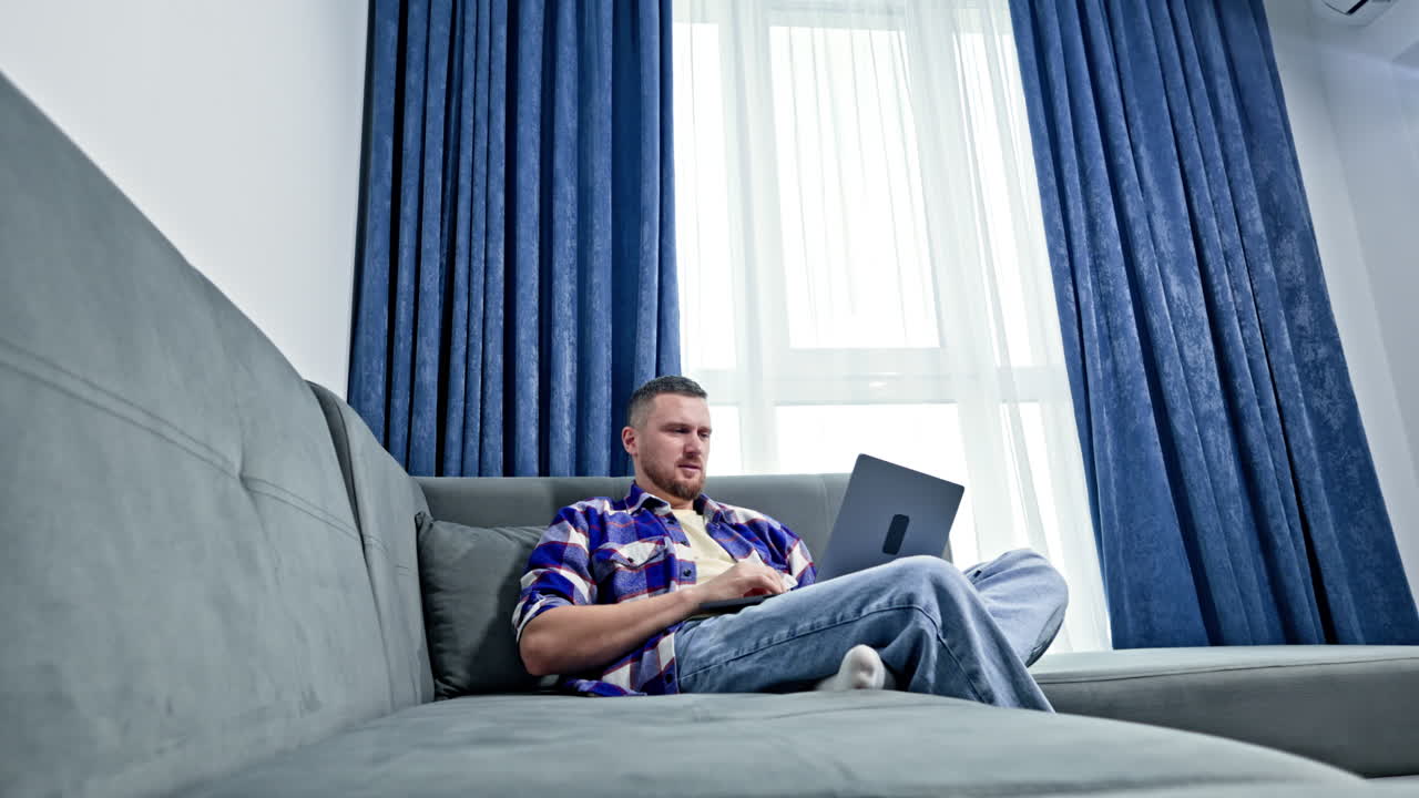 Man works on laptop while sitting on gray couch in modern living room with large window and blue curtains during daytime
