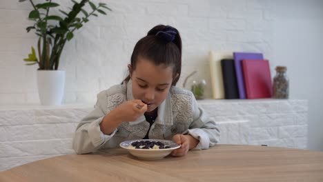 close-up of a cute baby girl with pleasure and appetite picks up raspberries and blueberries and puts them in her mouth at the table at home in the kitchen