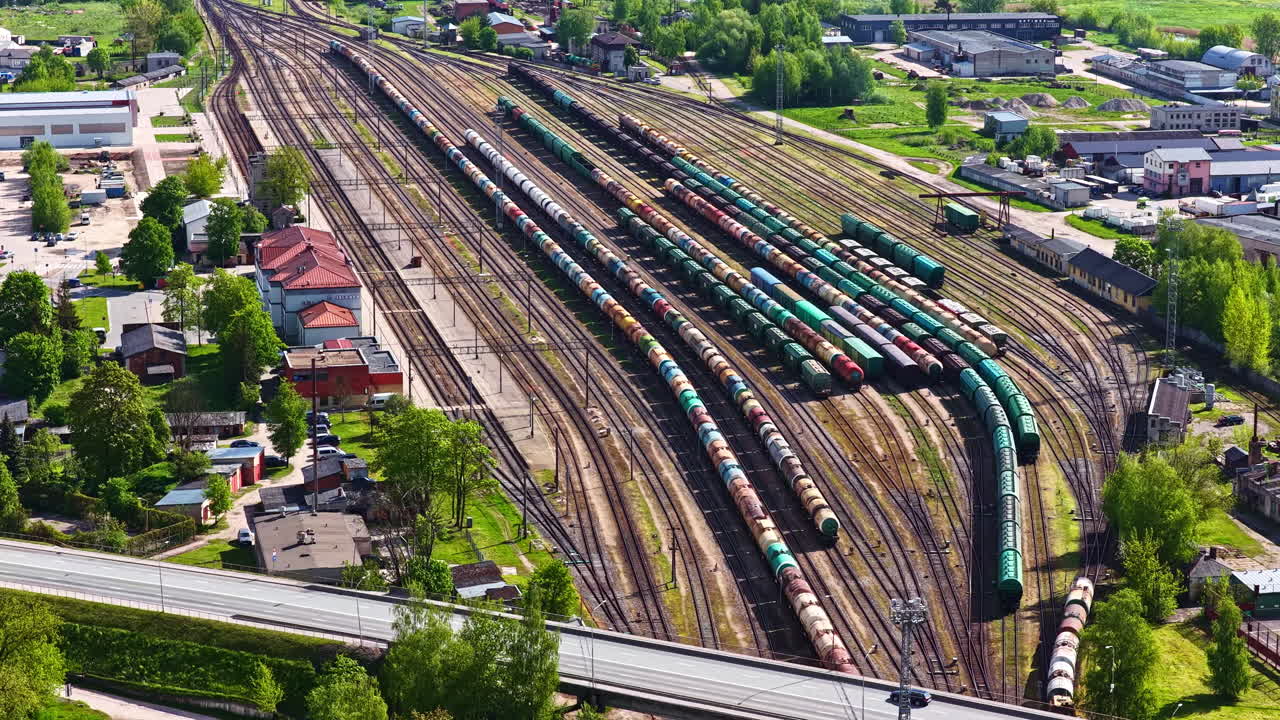 Luftfoto af en stor jernbaneplads med lange rækker af godsvogne, tankvogne og fragtlinjer, der strækker sig over et industriområde på en klar sommerdag.