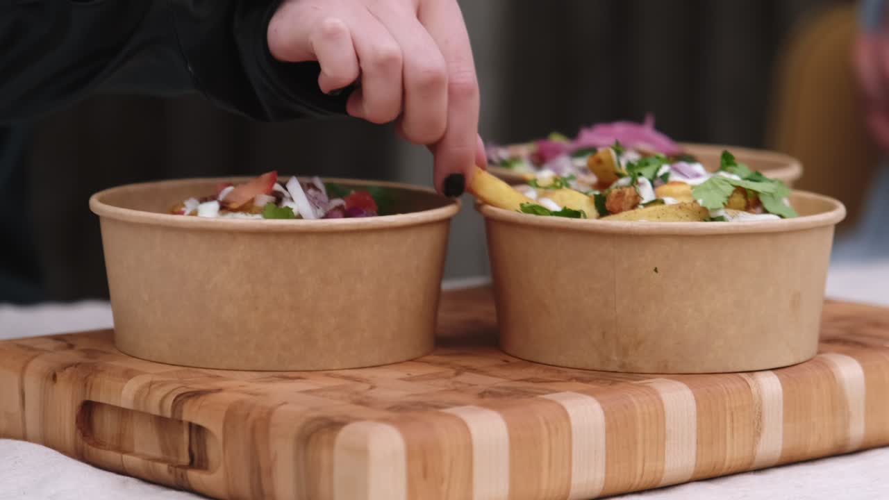 Close up of person picking up fries and eating Indian take away food from biodegradable poke bowl displayed on wooden tray.