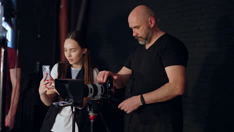 filming crew stands in the studio focused on recording video. male photographers and long-haired lady taking video on her phone laugh cheerfully.