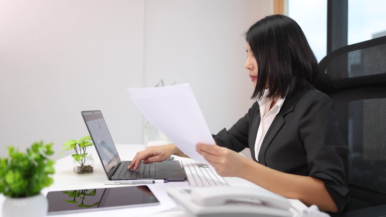Professional woman analyzes paperwork at desk, using laptop, in bright corporate workspace, static camera