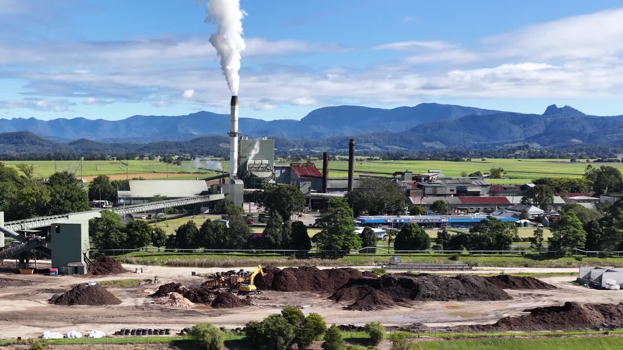 A sugar mill emits smoke against a backdrop of lush greenery and distant mountains under a clear blue sky