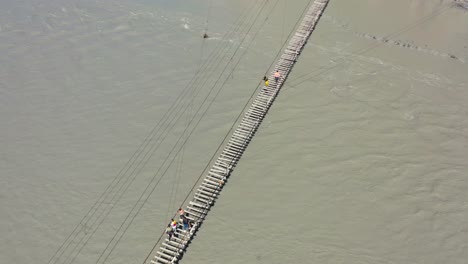 Vista-Aérea-De-Arriba-Hacia-Abajo-De-Los-Turistas-Cruzando-Con-Cautela-El-Famoso-Y-Peligroso-Puente-Hussaini-Sobre-Un-Río-Gris-De-Ritmo-Rápido-En-Hunza-Pakistán