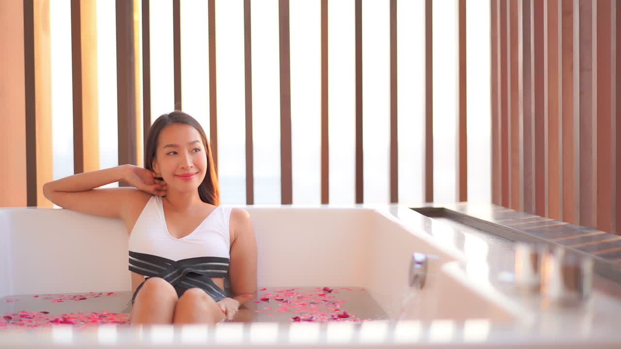 A young Asian woman, in a bathing suit, sits in a huge bath tube as it fills with water and rose petals
