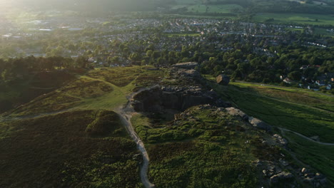 Establishing-Drone-Shot-with-Rays-of-Sun-of-Hangingstone-Rocks-Rock-Formation-on-Ilkley-Moor-with-Ilkley-Town-in-Valley-at-Golden-Hour-Sunset-UK