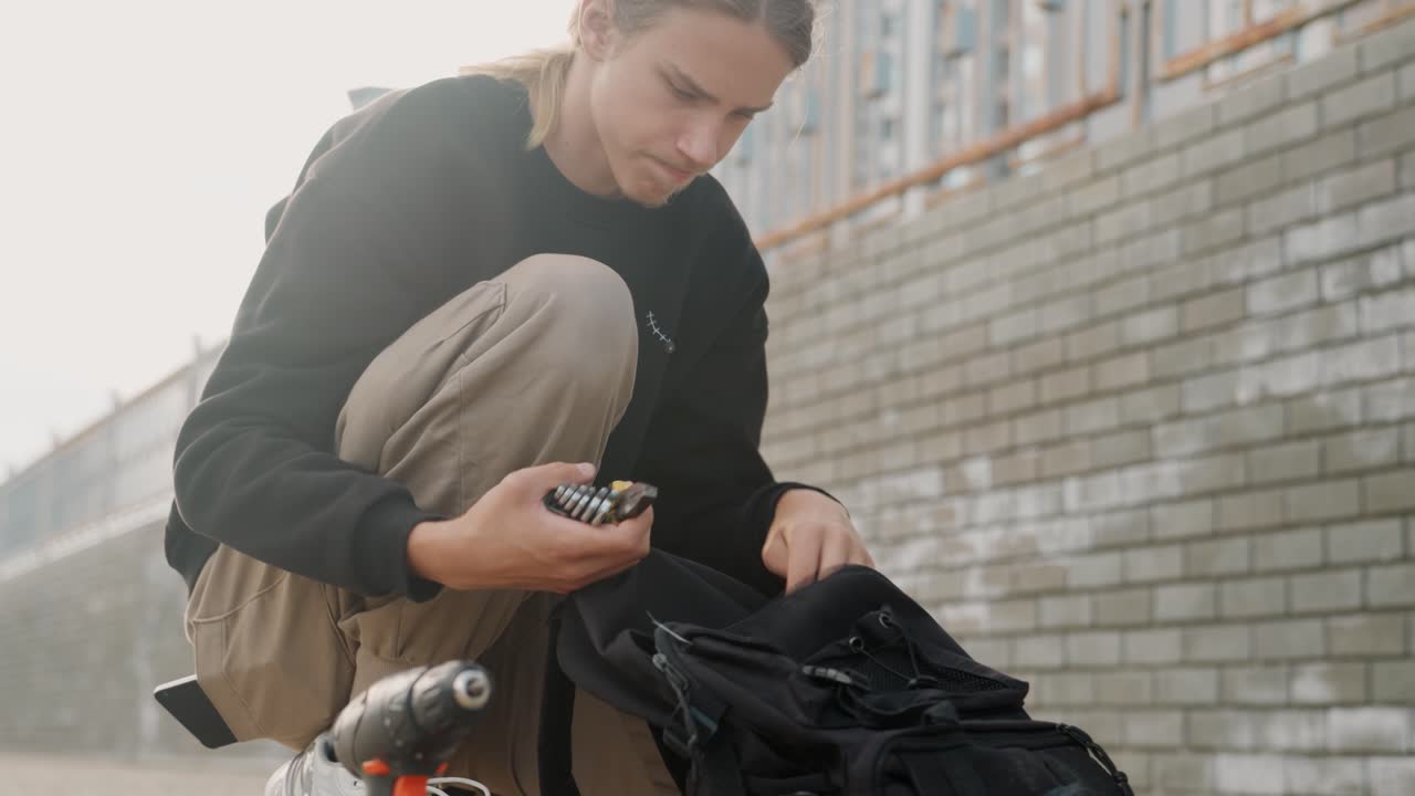 Young white courier checking phone, seated beside backpack and scooter at brickwall, inspecting small parts and messages, soft morning light, calm concentrated mood, casual cargo pants and hoodie