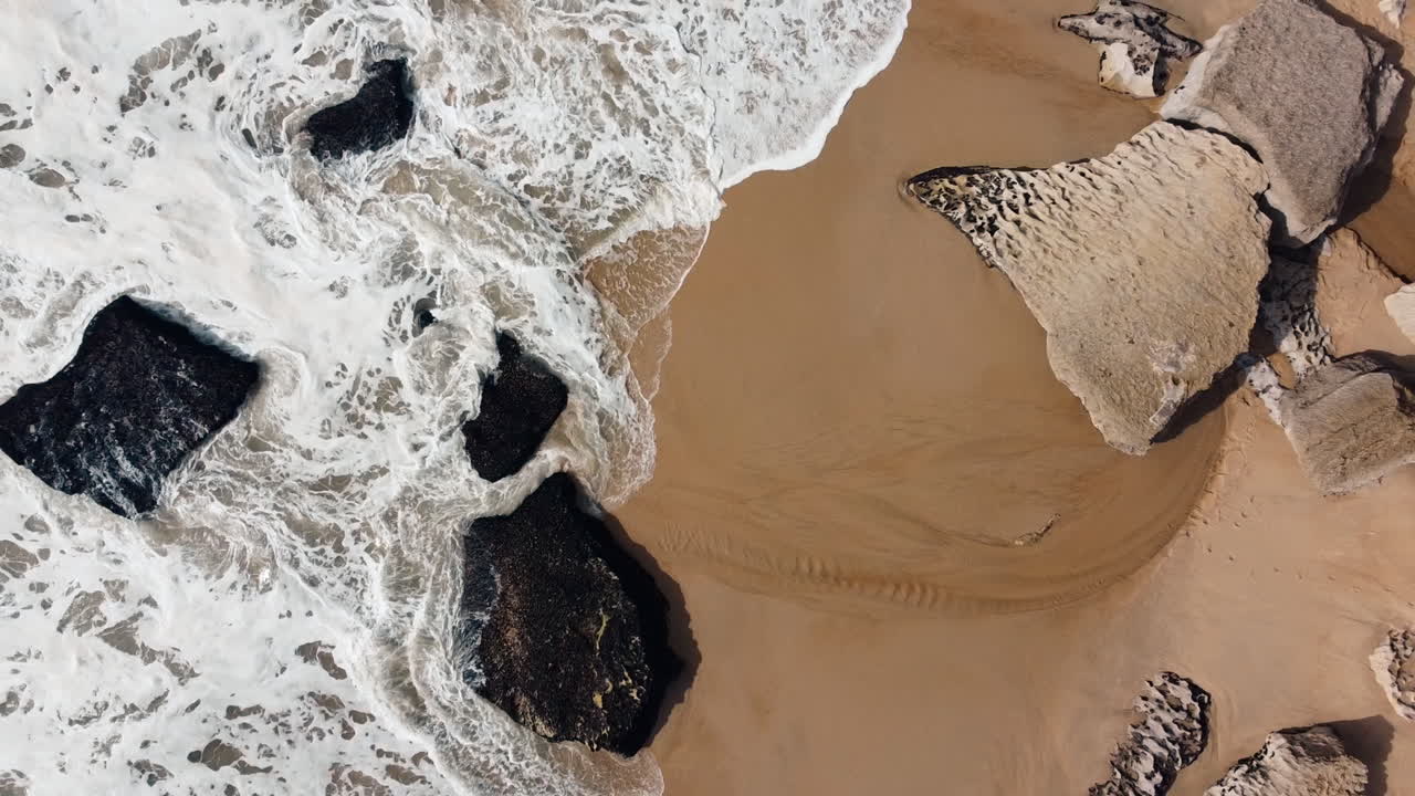 Bird-eye aerial of ocean waves crashing at rocky and stony beach in Nazare, Portugal