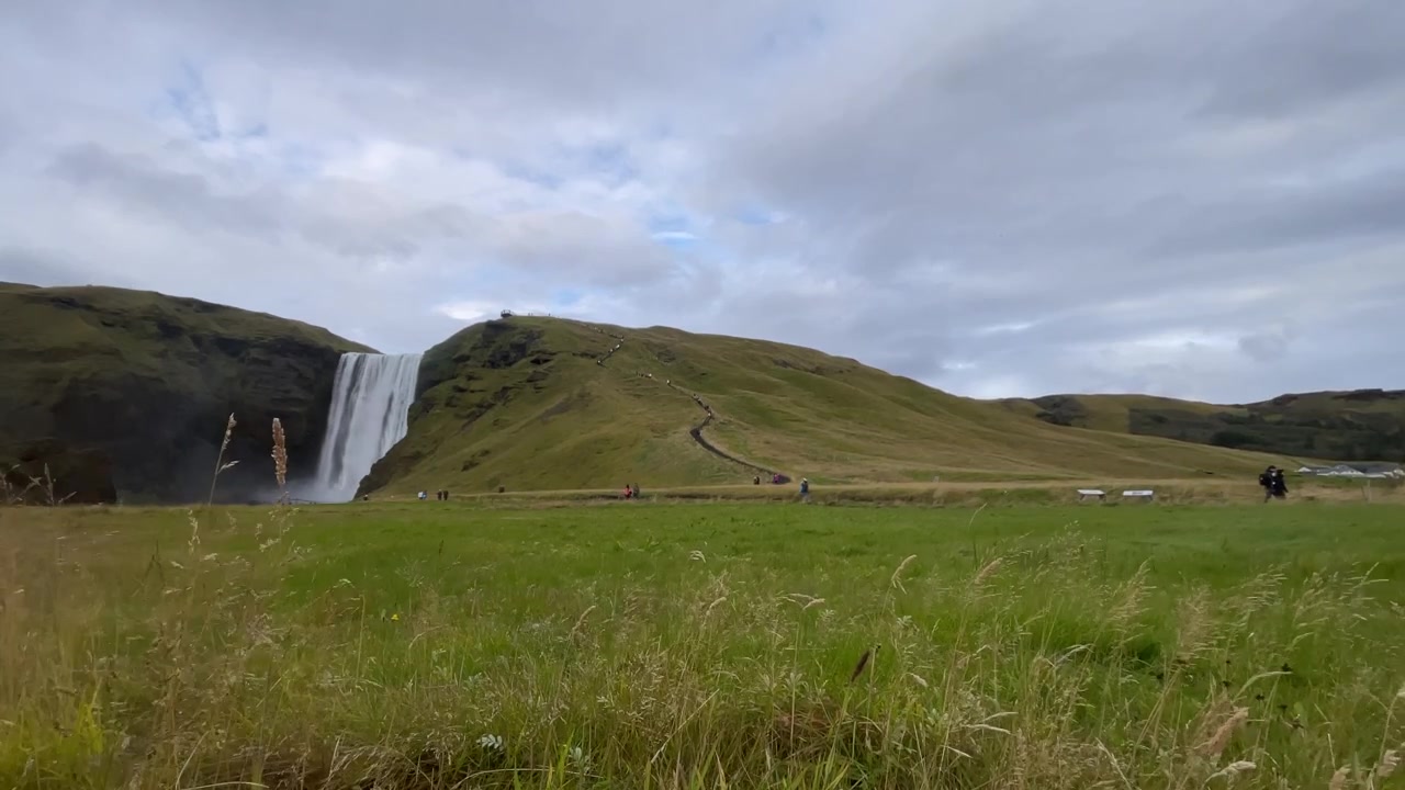 Ampia vista della cascata di Skogafoss circondata da rigogliose colline verdi e praterie nell'Islanda meridionale sotto un cielo nuvoloso