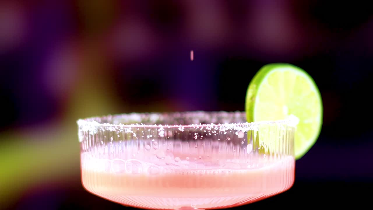 A vibrant pink liquid is poured into a salt-rimmed glass, garnished with a lime slice, against a blurred background.