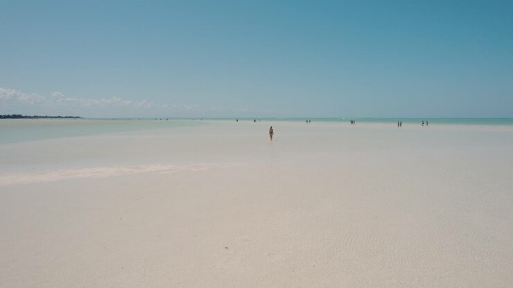 Attractive blonde girl in thongs walks on sandy beach with shallow sea on island paradise, reversing aerial