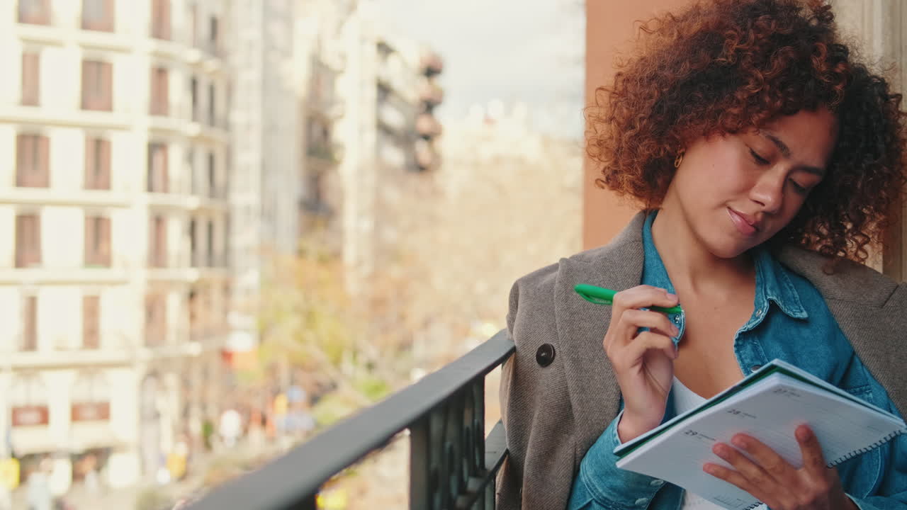 Thoughtful Young Woman Writing in Planner on Balcony