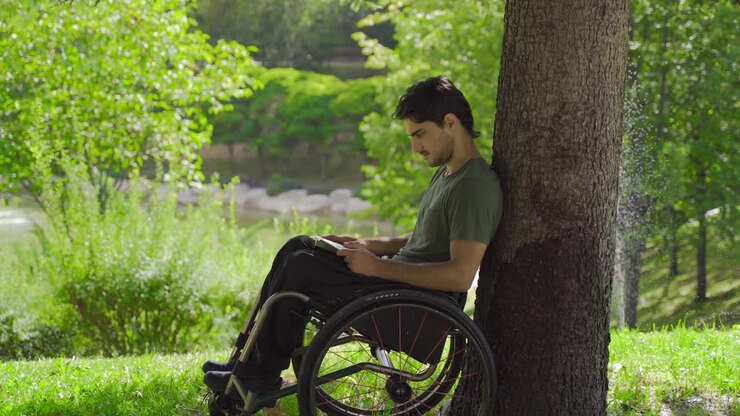 Teenager sitting in a wheelchair is reading a book.