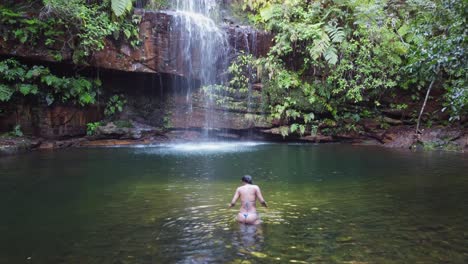 Frau-Im-Winzigen-Bikini-Betritt-Einen-Ruhigen-Pool-Unterhalb-Eines-Hübschen-Dschungelwasserfalls