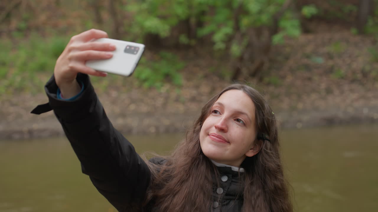 Mulher caucasiana posando para selfie perto de um rio, levanta o smartphone com um sorriso alegre, usando um casaco com capuz e cabelo castanho comprido, árvores verdes e margem do riacho ao fundo; os frames mostram caminhada, alcance, diversão