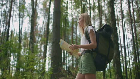 young caucasian woman looking for direction on a map while hiking in the forest. happy girl while hiking in nature and orienteering with help of a map