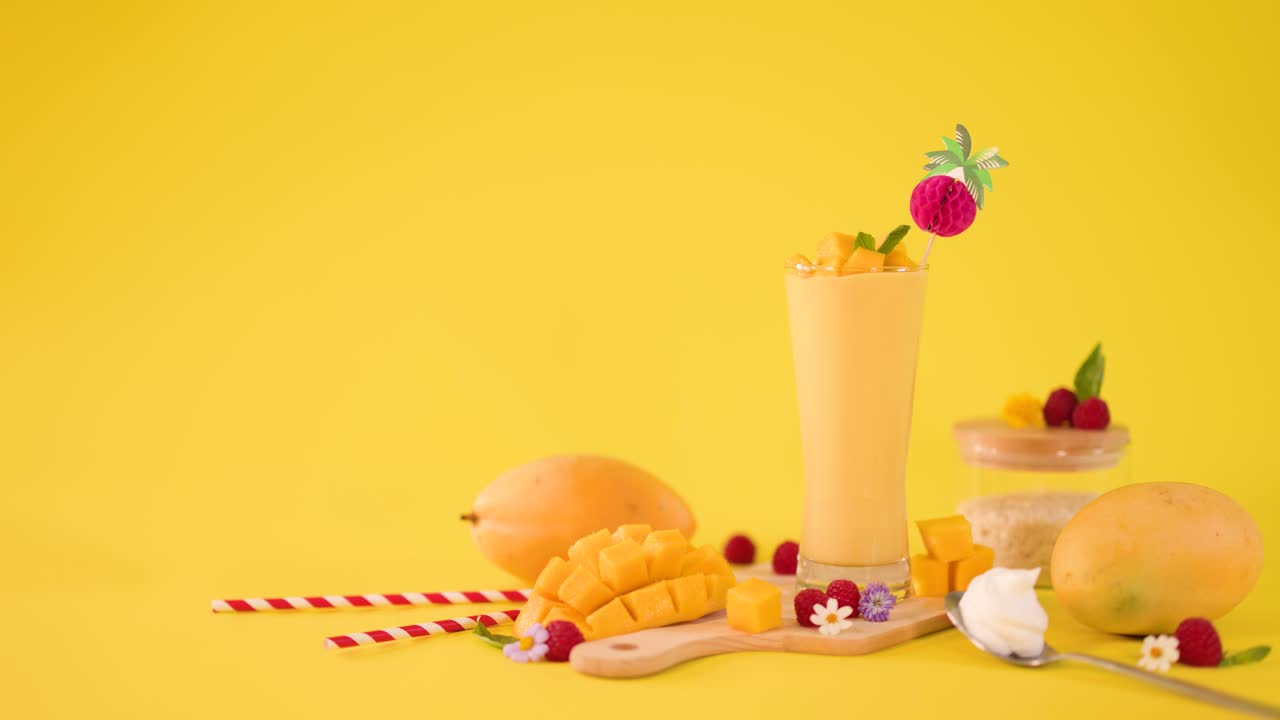 Una mano umana decora un frullato di mango alto con una cannuccia a righe e un lampone su uno sfondo giallo brillante
