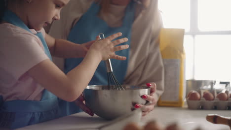 Una-Niña-Curiosa-Está-Ayudando-A-Mamá-En-La-Cocina-Mezclando-Masa-O-Crema-En-Un-Tazón-Con-Un-Batidor-Para-Cocinar-Pastel